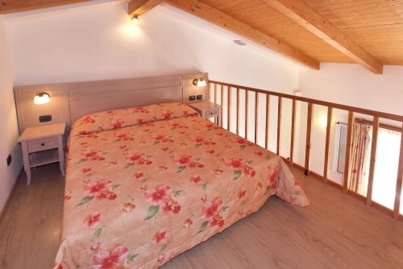 Une chambre avec un plafond en bois, un lit recouvert d’une couvre-lit à motifs floraux, une table de chevet et un luminaire fixé au mur.