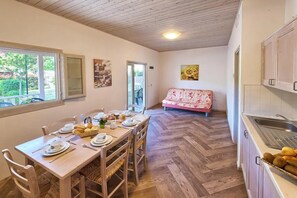 Un coin repas avec une table en bois mise pour le petit-déjeuner, un canapé en arrière-plan et une cuisine équipée d’un évier et de placards.