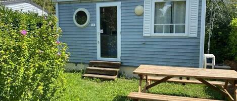 A small blue cabin with a front door, a round window, and a picnic table in the foreground.