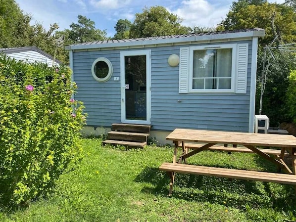 A small blue cabin with a front door, a round window, and a picnic table in the foreground.