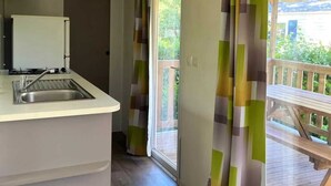 A compact kitchen with a sink and cabinets, a sliding glass door leading to a balcony, and a view of greenery outside. - ZEcamping 2* animaux acceptés dans un environnement calme et naturel (Angicourt)