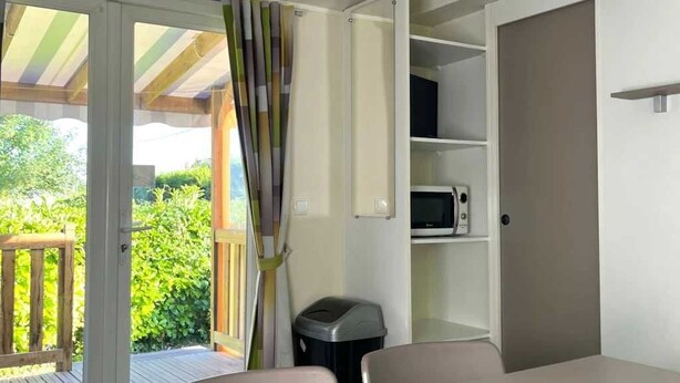 A modern room with a sliding glass door leading to an outdoor deck, a white table, and built-in shelving with a microwave. - ZEcamping 2* animaux acceptés dans un environnement calme et naturel (Angicourt)