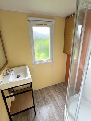 Ett litet badrum med ett vitt handfat, ett fönster och en duschhörna.