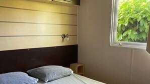 Et soveværelse med seng, puder, et natbord, et vindue med udsigt til grøn natur og indbyggede hylder.
