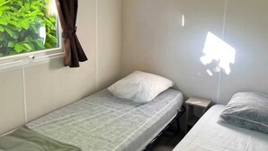 Un petit logement composé d’une seule pièce, avec deux lits simples, une fenêtre avec des rideaux et une table de chevet.