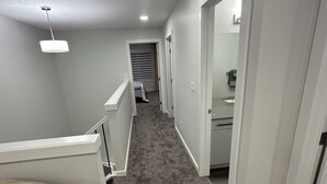 Un couloir étroit au sol recouvert de moquette, aux murs blancs et éclairé par un plafonnier. On aperçoit un lit sur la gauche et, sur la droite, une salle de bain à travers une porte ouverte.