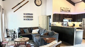 A modern living room with a grey sofa, a wooden coffee table, and a cowhide rug. There is a kitchen area with dark wooden cabinets and a stainless steel refrigerator. - 3B/2BA Cabin-Style Condo in Pinetop Lakes. Steps from Golf, 30 Mins to Sunrise! (Pinetop Country Club)