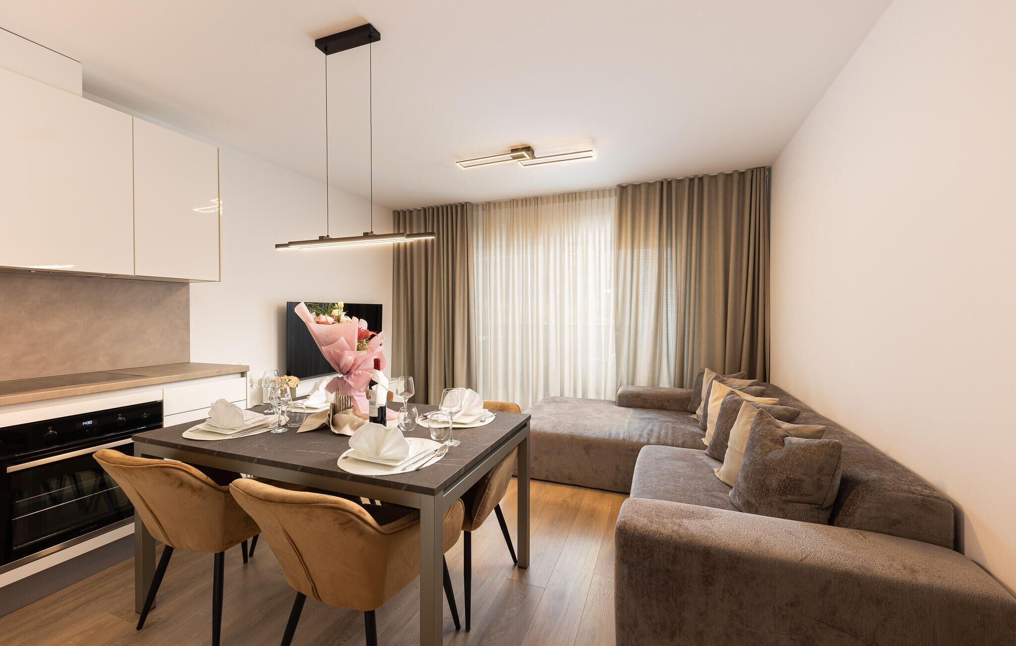 Un comedor moderno con una mesa de madera oscura dispuesta para dos personas, un sofá y un televisor al fondo.