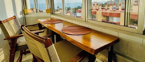 A dining area with a wooden table and chairs, large windows with a view of buildings and the sea.
