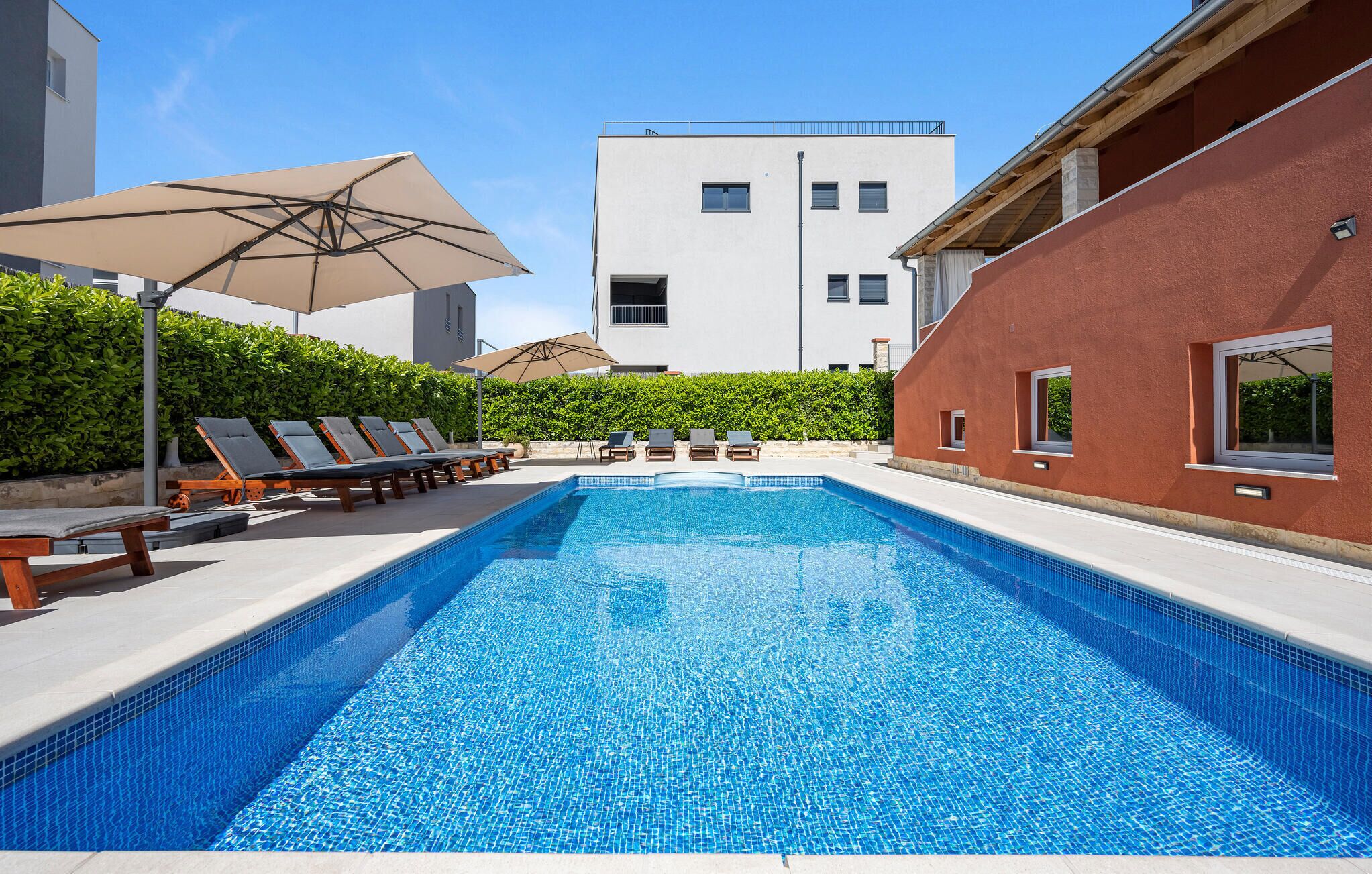 Una piscina con sillones de descanso y sombrillas, rodeada de edificios y vegetación.
