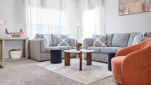 Een woonkamer met een grijze bank, een ronde salontafel en een oranje fauteuil. Er staat een klein tafeltje met een 'welcome'-bordje en een mand.