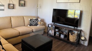 Una sala de estar con un sofá color beige, un reposapiés negro, un televisor sobre un mueble de madera y una pared con un letrero que dice 'be our guest'.