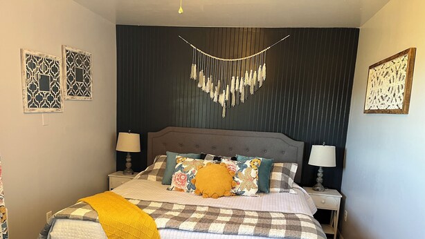 Una cama bien hecha con colcha acolchada, varios almohadones y una manta amarilla. Hay dos mesitas de noche con lámparas, y la habitación tiene una pared oscura con paneles decorativos.