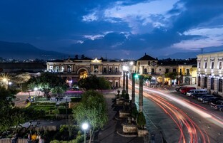 A historic building with classical architecture, a central fountain, and a well-lit street with light trails.