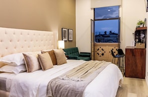 A hotel room with a large bed, a green armchair, a desk, and a view of the cityscape through a large window. - Central Park Jr. Suites (Quetzaltenango)