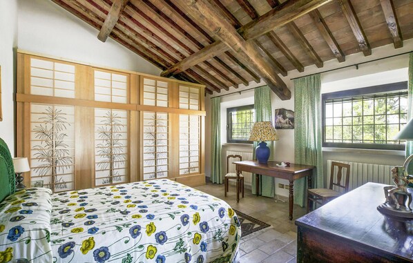 A traditional room with a wooden ceiling, a bed with a floral bedspread, a table with a lamp, and a window with curtains.