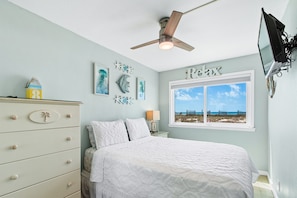 A bedroom with a bed, a ceiling fan, a dresser, a window with a view, and wall decorations.