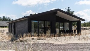 A modern, black, single-story building with large glass windows, situated on a gravel and sand terrain with sparse vegetation. - Stunning home in Oksbøl with sauna (Oksbøl)