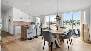 A modern dining area with a wooden table and grey chairs, large windows with a view of the outdoors, and a kitchen area with a wooden countertop and bar stools. - Stunning home in Oksbøl with sauna (Oksbøl)