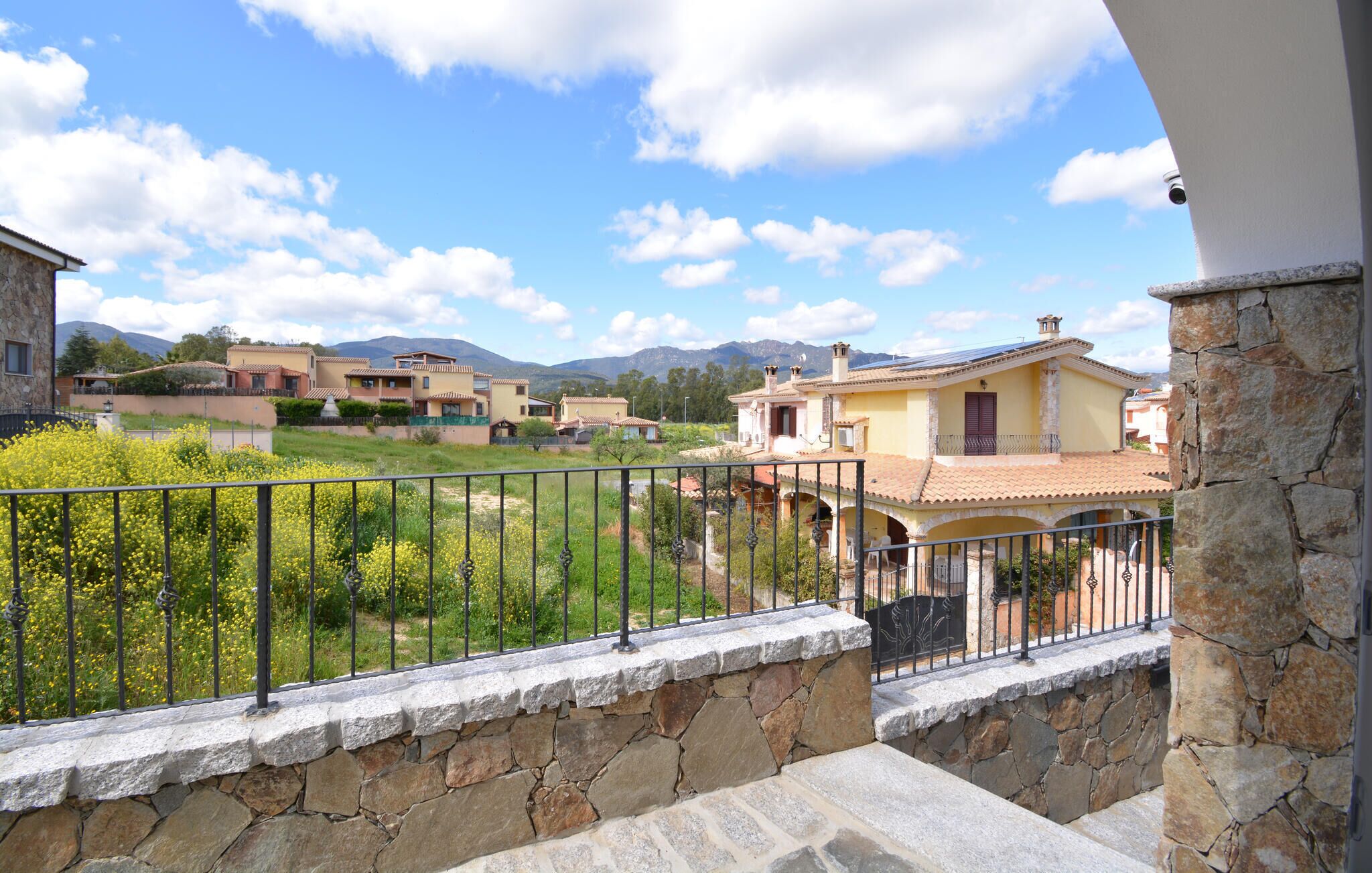 Un balcón con pared de piedra y barandilla de metal, con vistas a una zona residencial con casas y montañas al fondo.