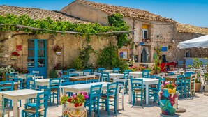 Un café en plein air avec des chaises bleues et des tables blanches, entouré de bâtiments en pierre aux toits de tuiles et à la végétation luxuriante.