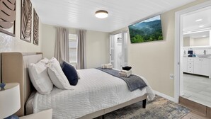 Ein Schlafzimmer mit Bett, Nachttisch, Lampe, einem Fenster mit Vorhängen, einem Bild von einem Wald und einem durchgegangenen Tür sichtbaren Badezimmer.