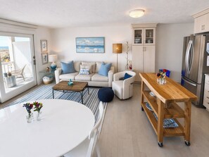 Una sala de estar moderna con sofá, mesa de centro y mesa de comedor, una cocina con refrigerador, y un balcón con una silla.