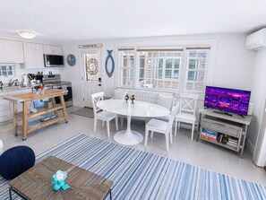 Una cocina moderna con armarios blancos, un comedor con mesa y sillas blancas, un televisor sobre un mueble bajo y un ventanal grande.
