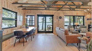 Interior de una cabaña de madera con zona de comedor, sofá y asientos junto a un gran ventanal con vistas a un lago.