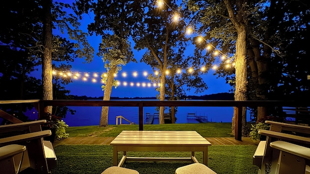 Una terraza al aire libre con una mesa y bancos de madera, rodeada de árboles y luces de cuerda, con vistas a un cuerpo de agua.