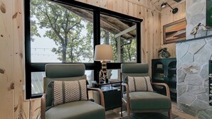 Una habitación acogedora con dos sillones verdes, una mesa con lámpara, una chimenea de piedra y un gran ventanal con vistas a árboles.