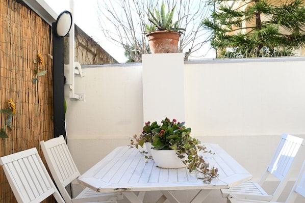 A white outdoor table with a potted plant, two white chairs, and a bamboo wall.