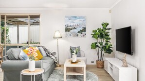 Una sala de estar moderna con un sofá gris, una mesita de centro, una lámpara de pie, una planta verde grande y un televisor de pantalla plana montado en la pared.