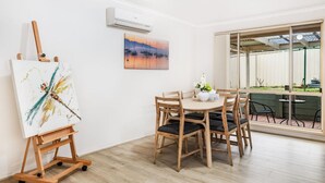 Una habitación con una mesa de comedor de madera, sillas, un caballete con una pintura, aire acondicionado y una ventana con vistas al exterior.