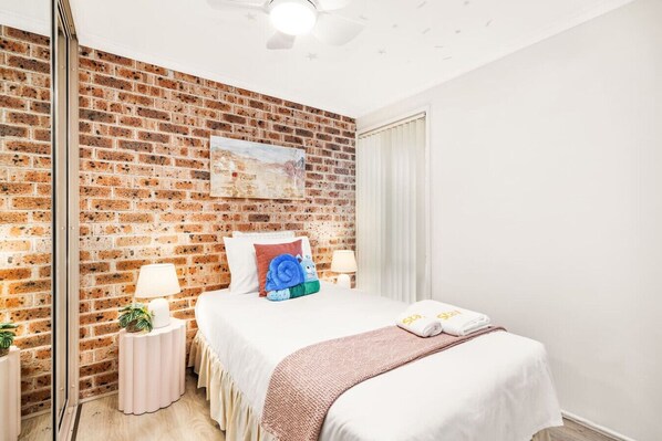 A hotel room with a brick wall, a bed with white bedding, two bedside tables with lamps, and a ceiling fan.