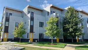 A modern multi-story residential building with a flat roof, large windows, and a small front yard with a picnic table.