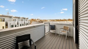 A rooftop terrace with a view of the city skyline, featuring a wooden deck, metal railings, and two chairs.