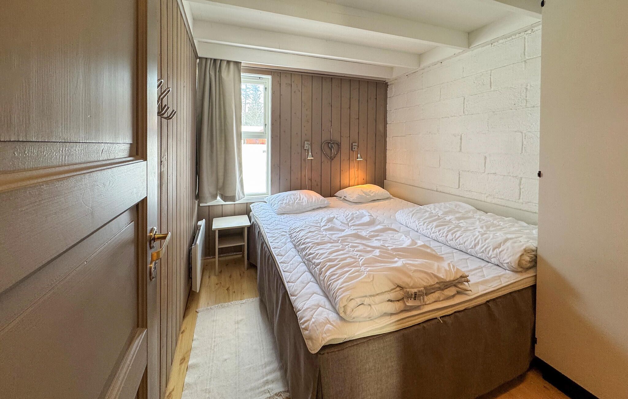 A bedroom with a wooden door, a window with curtains, a bed with white bedding, and a white brick wall.