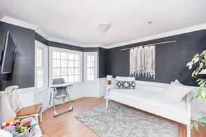 Una habitación con una cama blanca, una alfombra gris, una silla blanca, una mesita pequeña y un colgador de macramé en la pared.