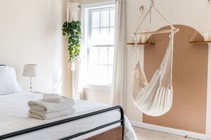 A bedroom with a bed, a hanging plant, a hammock, and a window with curtains.