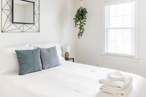 Una cama bien hecha con sábanas blancas, dos almohadas y una pila de toallas dobladas. Un espejo geométrico cuelga de la pared, y se observa una planta colgante.