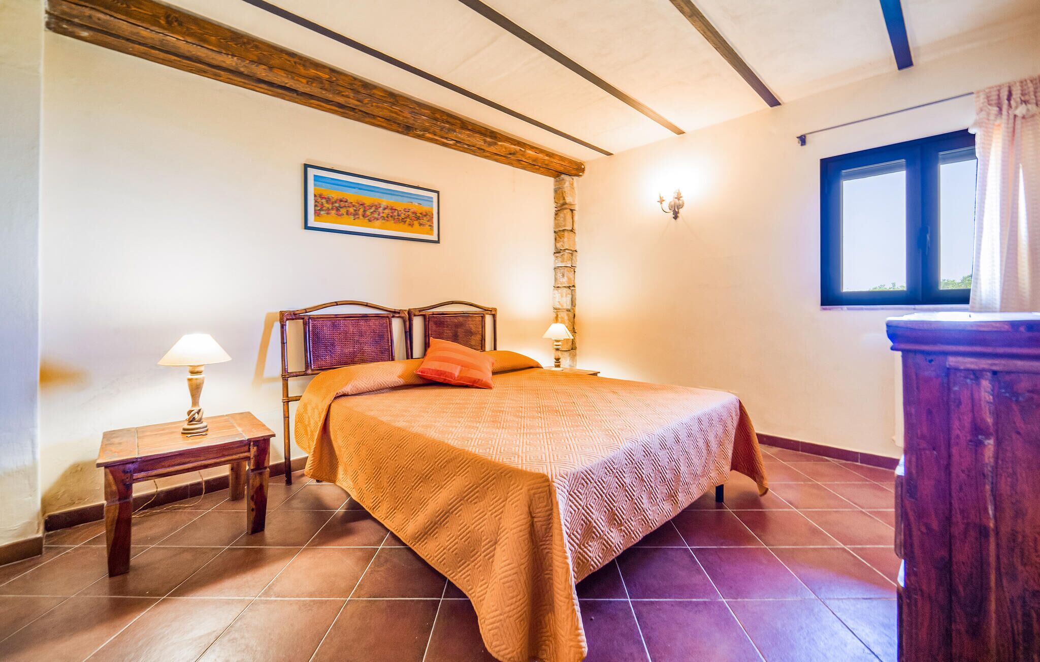 Un dormitorio con una cama, una mesita de noche, una lámpara, un muro de piedra, una ventana y un cuadro en la pared.