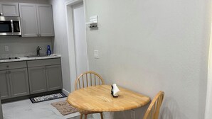 Eine kleine Küche mit grauen Schränken, einer Mikrowelle und einem Spülbecken. Ein Esstisch mit zwei Stühlen und einem Teppichboden sind ebenfalls vorhanden.