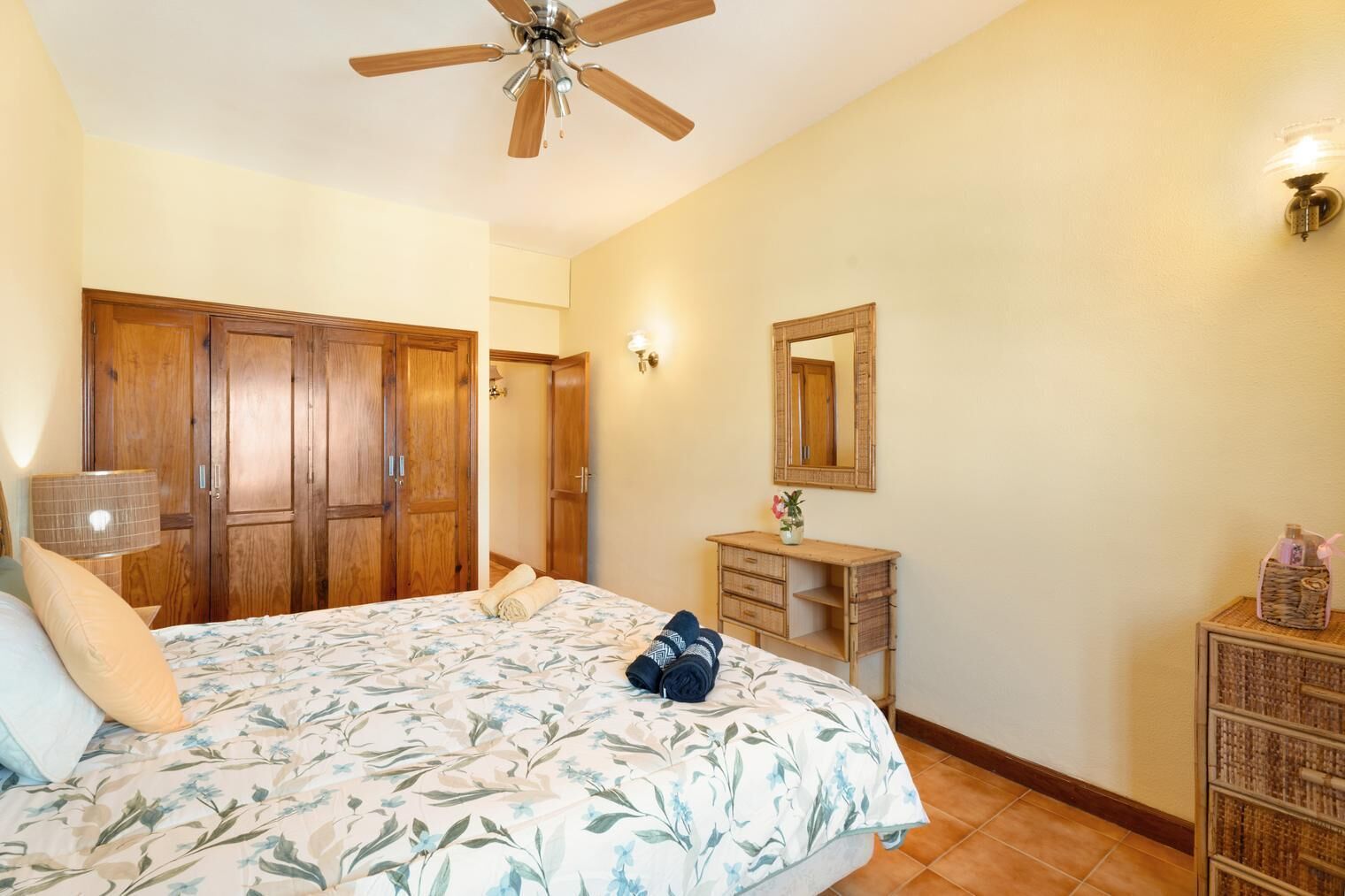 A bedroom with a bed, wooden wardrobe, bedside table, mirror, and a ceiling fan.