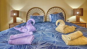 A double bed with a blue leaf-patterned bedspread, two headboards, two bedside lamps, and folded towels in purple and beige.