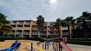 Un espace de jeux pour enfants avec un toboggan et des balançoires, entouré de palmiers et bordé d’un bâtiment doté de balcons.