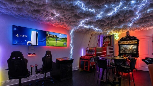 Una sala de juegos con múltiples pantallas, sillas gamer y techo iluminado con neón.