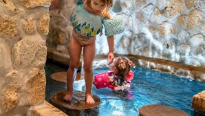 Un niño parado sobre un borde de piedra en una zona de piscina, con otro niño en el agua.