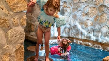 Un enfant se tient debout sur une bordure en pierre dans une zone de piscine, tandis qu’un autre enfant se trouve dans l’eau.