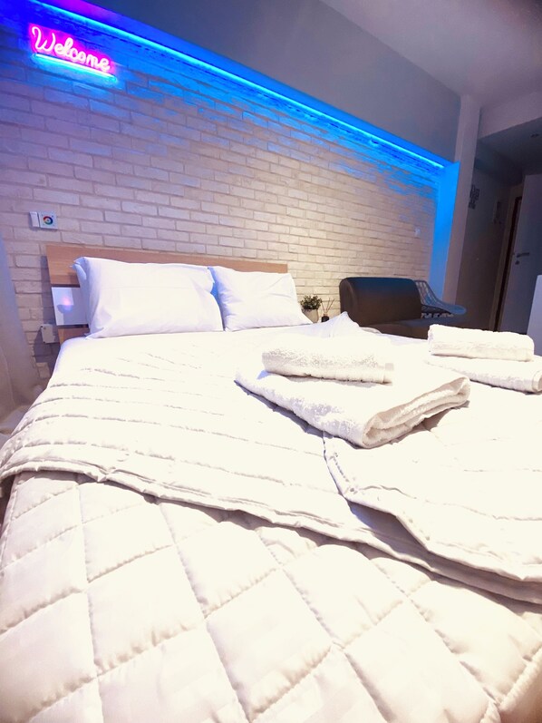 Habitación de hotel con pared de ladrillo, una cama con ropa blanca, un letrero de neón que dice 'Welcome' y una silla.